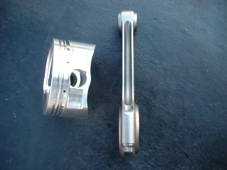 Aluminum piston and connecting rod on dark blue surface. "SUPERMOTORS www.supermotors.net" text in lower right