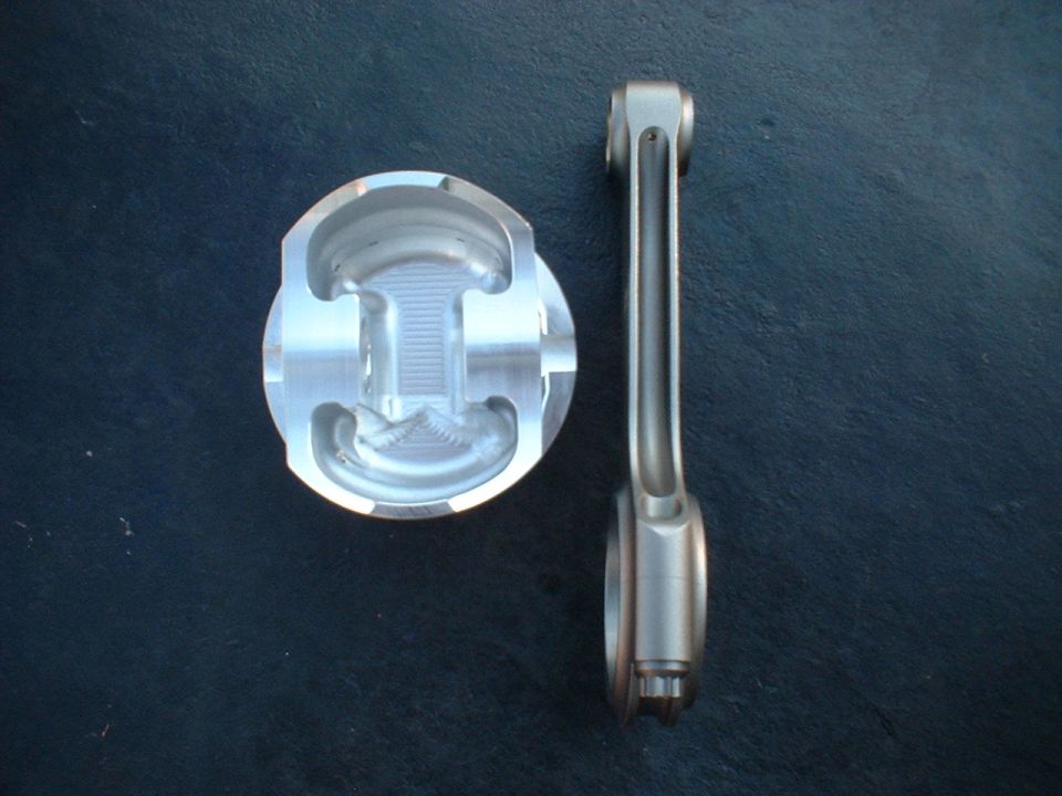 Aluminum piston and connecting rod on dark surface. "SUPERMOTORS www.supermotors.net" text visible