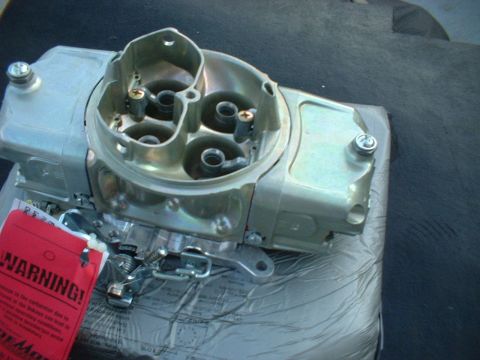 1968 Ford Galaxie - A shiny metal carburetor sits on a surface, with a red warning tag attached to its side.