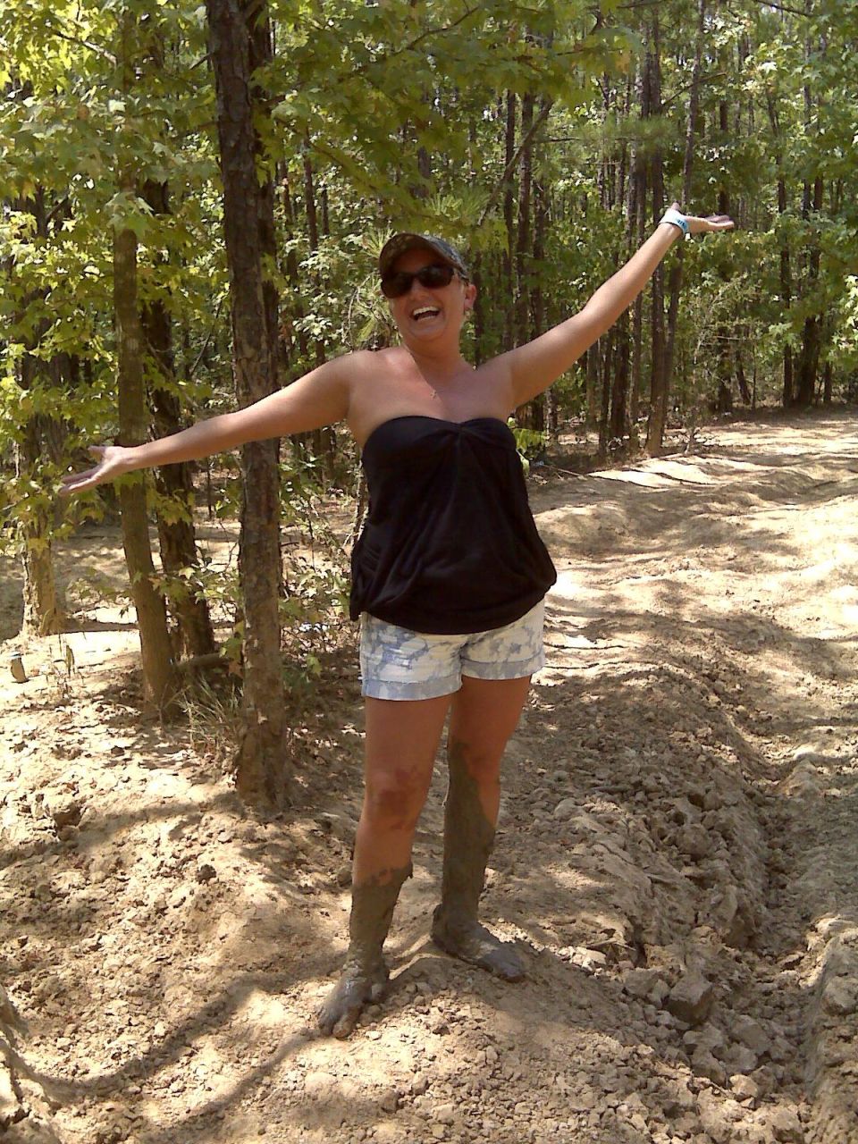Woman with arms outstretched in a wooded area, wearing a black strapless top, patterned shorts, and muddy boots
