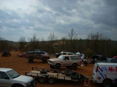 1996 Ford Bronco - A gathering of trucks and SUVs on a dirt lot with trees in the background and a cloudy sky overhead.