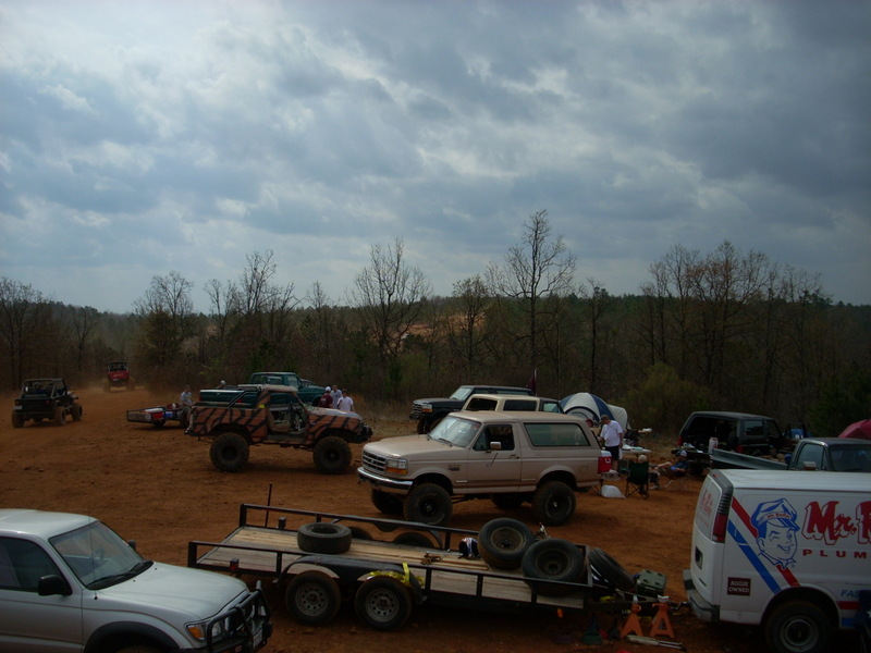 Several off-road vehicles and trailers parked on a dirt lot under a cloudy sky. One vehicle is a tan Ford Bronco