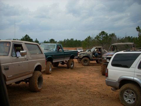 1996 Ford Bronco - Several off-road vehicles are gathered on a dirt lot under a cloudy sky.