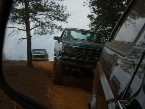1996 Ford Bronco - Two pickup trucks driving on a dirt road with trees and cloudy sky overhead.