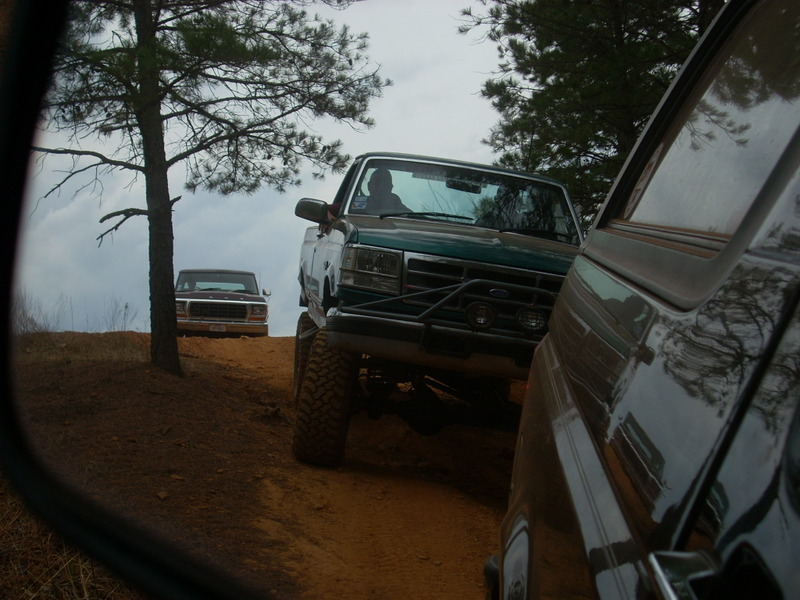 Green pickup truck with aftermarket bumper and large tires on a dirt trail, another pickup truck in the background