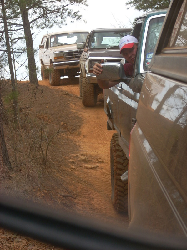 Several lifted trucks and SUVs driving single file on a dirt trail. A guy in a white and red hat is looking out the window of the closest truck