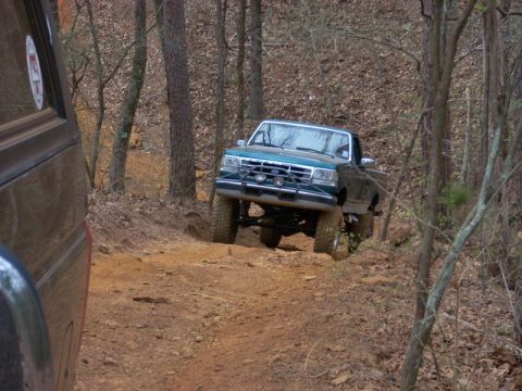 1996 Ford Bronco - A green 1996 Ford Bronco is seen off-roading on a dirt path, with another vehicle partially visible in the foreground.