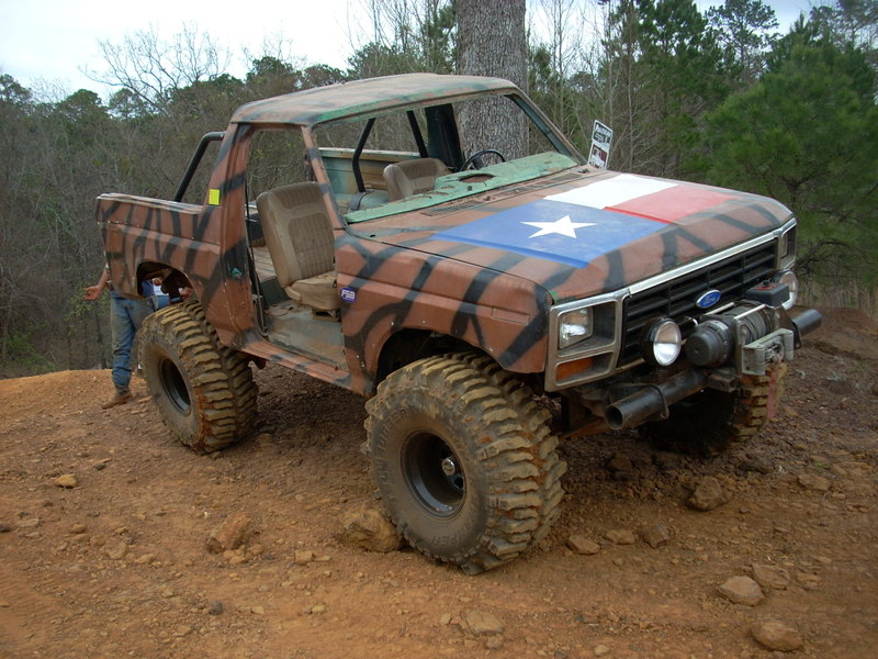 Modified off-road SUV with camouflage paint and Texas flag on hood, large tires, winch, and roll cage