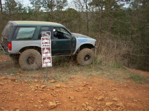 1996 Ford Bronco - Modified SUV with large tires navigates a wooded trail next to a trail sign.