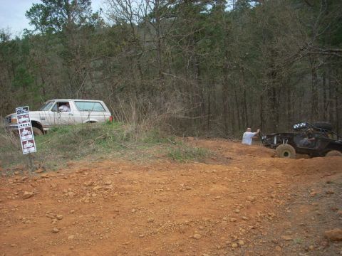 1996 Ford Bronco - Two off-road vehicles navigate a wooded trail with red dirt and sparse vegetation.