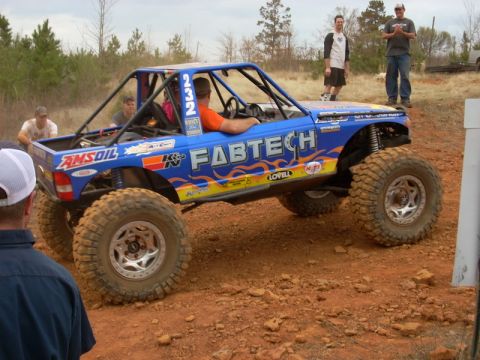 1996 Ford Bronco - A heavily modified blue off-road truck with a roll cage and large tires navigates a dirt track.
