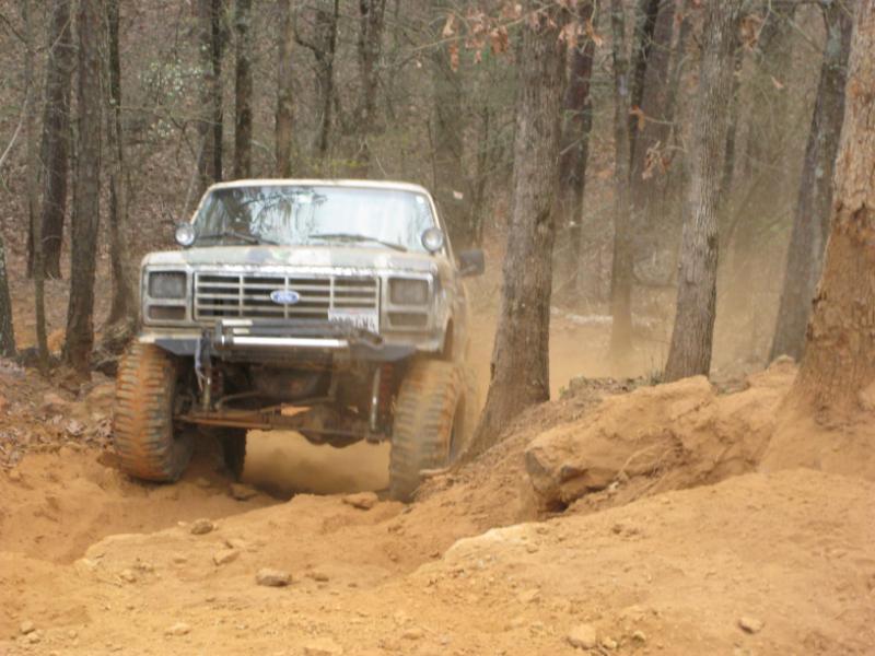 Ford Bronco driving on a dirt trail in woods. It has a camo paint job and large tires