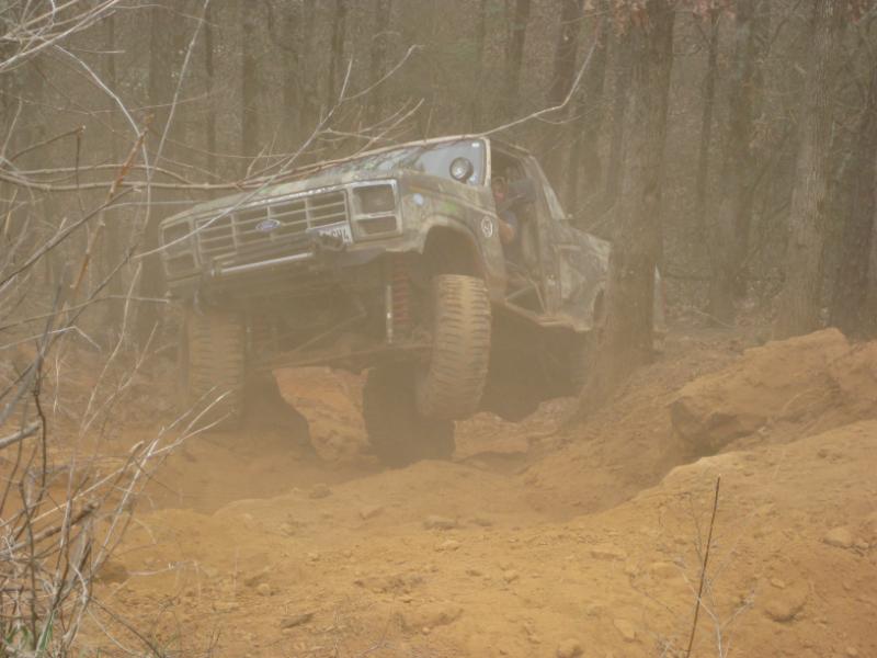 Modified off-road truck with large tires climbing a steep dirt incline, kicking up dust