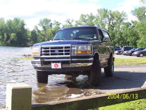 1990 Ford Bronco - A blue Ford Bronco is parked with its front tires in the water, with a parking lot in the background.
