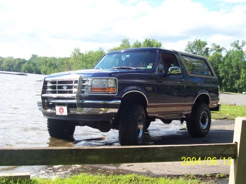 1990 Ford Bronco - A lifted Ford Bronco sits partially in the water, with trees and a cloudy sky in the background.