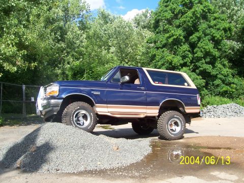 1990 Ford Bronco - A lifted blue Ford Bronco with tan trim is shown driving over a pile of gravel.