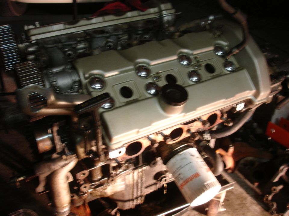 Engine block with a white oil filter attached. Some exhaust manifold pipes are visible