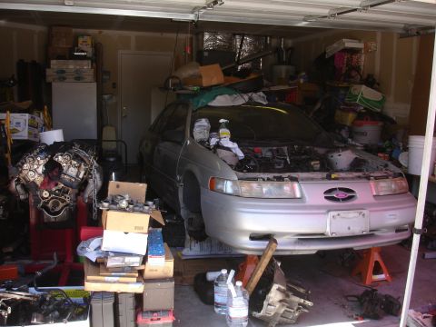 1992 Ford Taurus - A silver 1992 Ford Taurus sits in a garage with its hood open and engine removed, surrounded by car parts and storage.