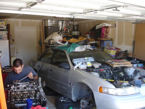 1992 Ford Taurus - A man works on the engine of a silver 1992 Ford Taurus in a cluttered garage, with the hood open and parts spread around.