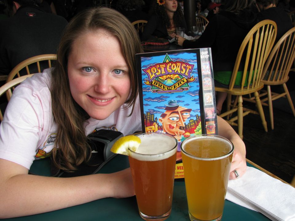 Woman smiles at the camera with two glasses of beer and a menu from Lost Coast Brewery