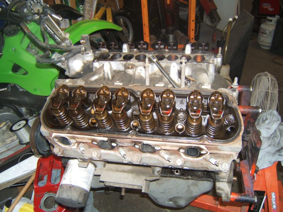 V8 engine block with valve covers removed, showing rocker arms and valve springs. A green dirt bike is partially visible in the background