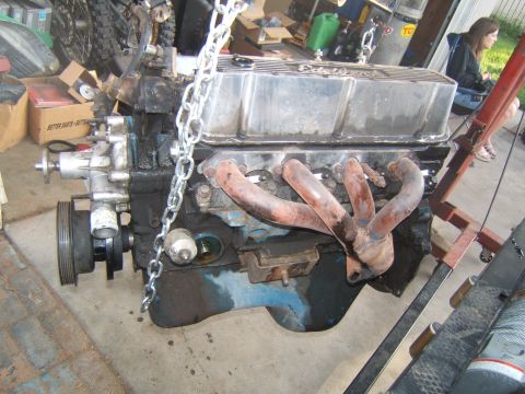 1988 Ford Bronco - Engine block with exhaust manifold and valve cover, suspended by a chain in a garage setting.