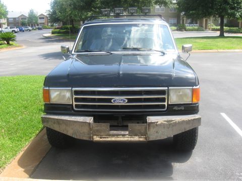 1990 Ford Bronco - Head-on view of a black Ford Bronco parked in an apartment lot, with a bare fabricated steel front bumper and winch cradle, bar grille with…