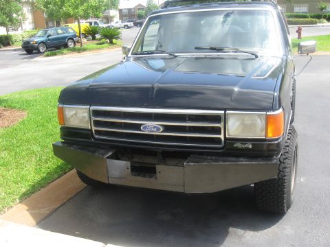 1990 Ford Bronco - Front 3/4 view of a black Ford Bronco parked on an apartment-complex driveway, wearing a bare unpainted fabricated steel front bumper, fact…