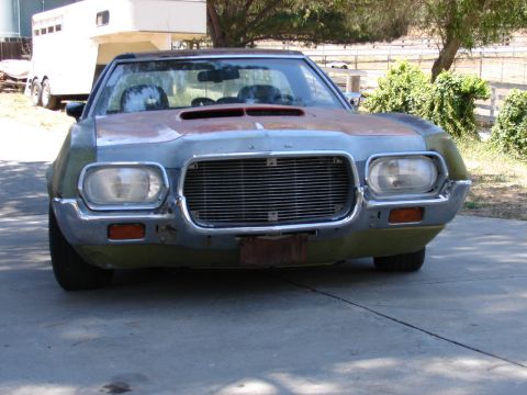1973 Ford Ranchero - Head-on view of a 1970s Ford car-based pickup wearing patchy green paint with bare primer and body filler across the hood and front fenders…