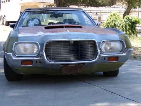 1973 Ford Ranchero - Head-on view of an unfinished early-1970s Ford Ranchero/Torino-style front end sitting on a concrete driveway, with faded olive-green paint…