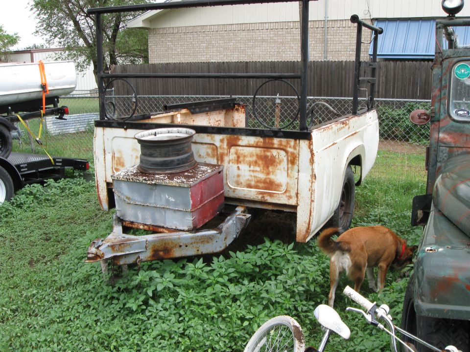 Rusty white pickup truck bed with a spare tire and toolbox on top, parked next to a green vehicle. A dog sniffs near the green vehicle