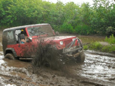 1979 Chevrolet Blazer - A red Jeep Wrangler navigates through a muddy off-road trail, kicking up a large spray of water.