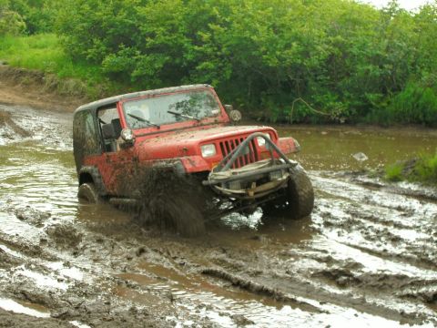 1979 Chevrolet Blazer - A red Jeep Wrangler navigates a muddy off-road trail, kicking up mud as it drives through a water-filled rut.