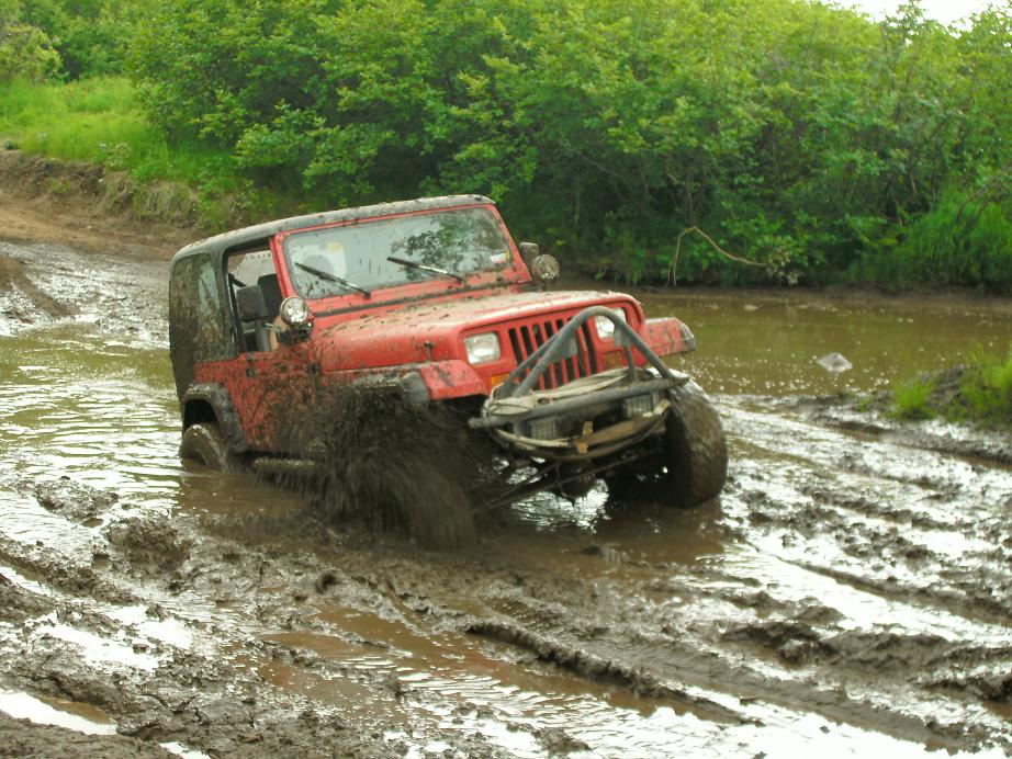 Red Jeep Wrangler driving through mud with front bumper and tires covered in mud
