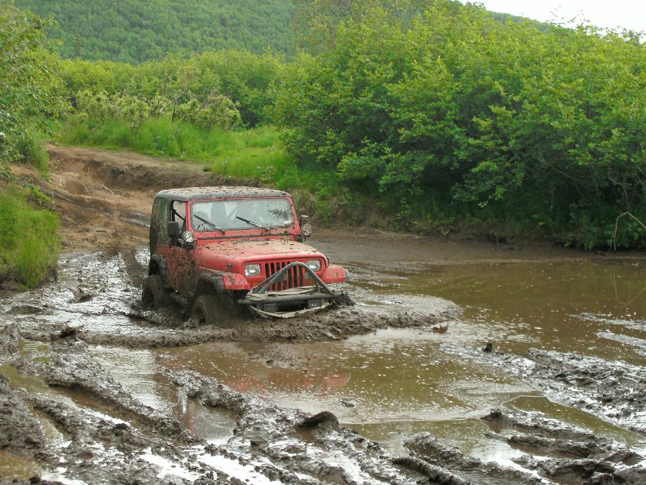 Red Jeep Wrangler deep in a mud pit on a trail, front bumper submerged
