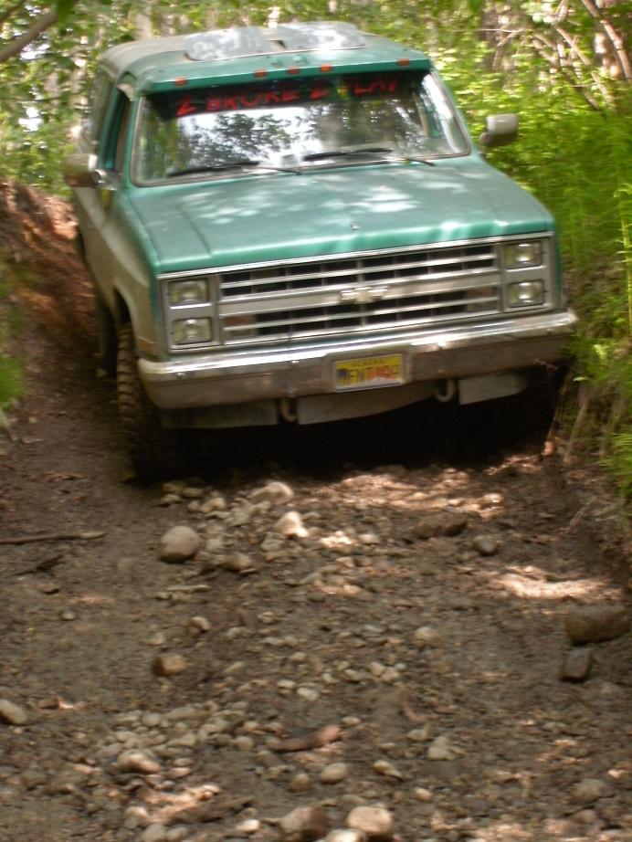 Green Chevrolet K5 Blazer driving on a rocky, wooded trail. Sunroof visible