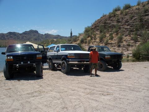 1996 Ford Bronco - Three SUVs are parked on a sandy shore near a lake, with a man standing between two of them.