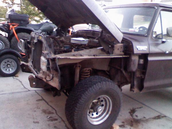 Brown Ford Bronco with hood up and front clip removed, exposing frame and coil spring in driveway