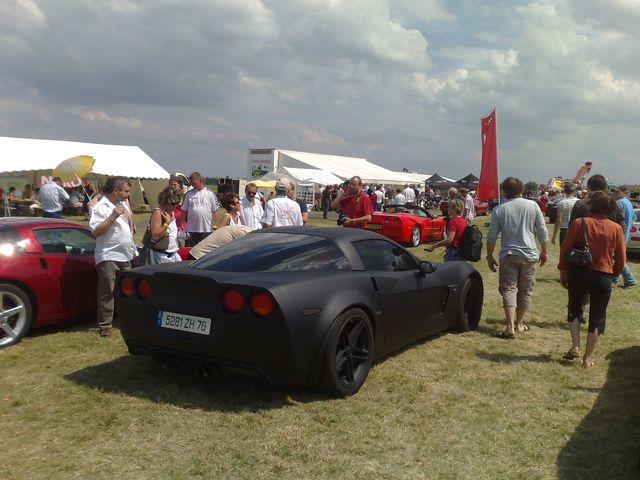 Black Chevrolet Corvette C6 parked on grass with people around. Red Corvette visible to the left