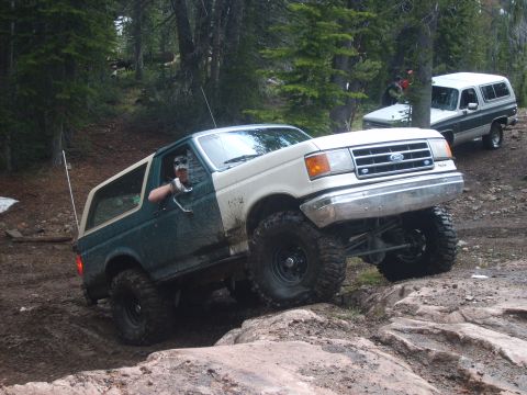 1989 Ford Bronco - Two SUVs navigate a rocky, wooded trail with one vehicle climbing over large stones.