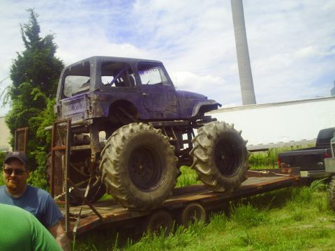 1990 Ford Bronco - A heavily modified purple monster truck with oversized tires is loaded onto a trailer, with a black pickup truck visible behind it.