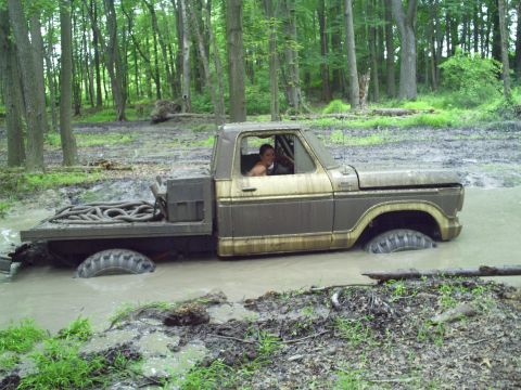 1990 Ford Bronco - A brown flatbed truck is deeply stuck in a muddy, wooded area, with a person visible in the driver's seat.