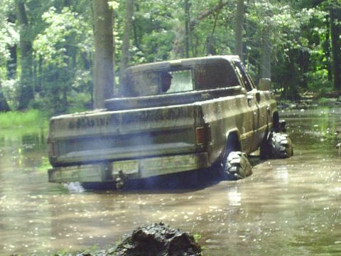 1990 Ford Bronco - A heavily soiled pickup truck is submerged in mud and water, with trees in the background.