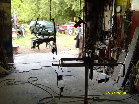 1989 Ford Bronco - A metal frame is suspended by chains in a workshop, with vehicles visible through the open garage door.