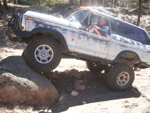 1978 Ford Bronco - A driver navigates a white 1978 Ford Bronco over a large rock on a dirt trail, demonstrating its off-road capabilities.