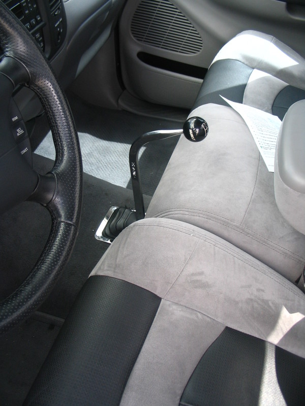 Aftermarket Hurst shifter with a black ball knob in a truck's center console, next to a perforated leather steering wheel