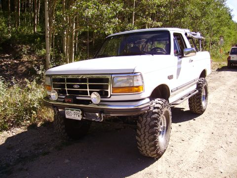 1978 Ford Bronco - A lifted white Ford Bronco is parked on a dirt road with trees in the background.