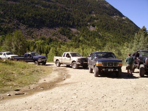 1978 Ford Bronco - A convoy of trucks and an SUV are stopped on a dirt road, with a dense forest and mountain visible behind them.