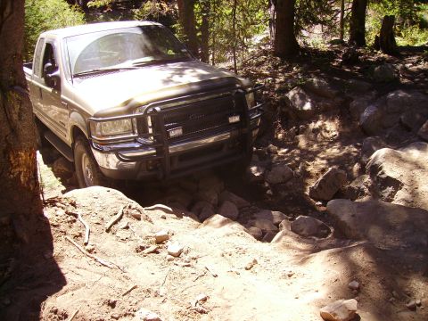 1978 Ford Bronco - A tan pickup truck navigates a challenging rocky trail through a wooded area.