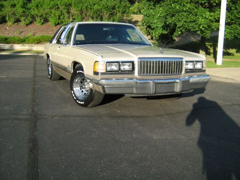 1988 Mercury Grand Marquis - A tan 1988 Mercury Grand Marquis sedan is parked on asphalt, viewed from the front with a prominent chrome grille.